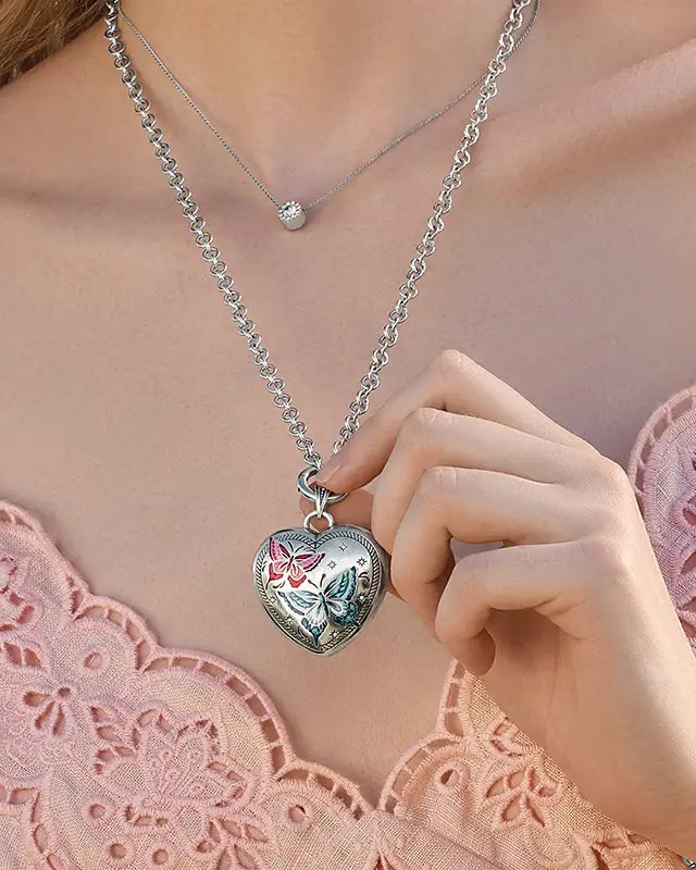Brighton Heart-shaped silver locket with floral design on a chain, held by a hand against a pink lace background.