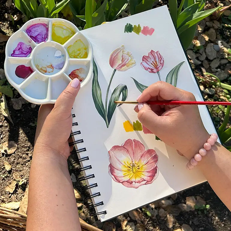 Person painting flowers in a notebook with a color palette outdoors.