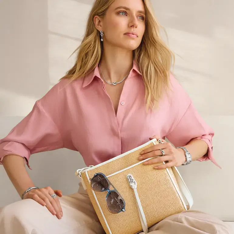 Woman in pink shirt holding a Brighton beige clutch with sunglasses, wearing jewelry, on a neutral background