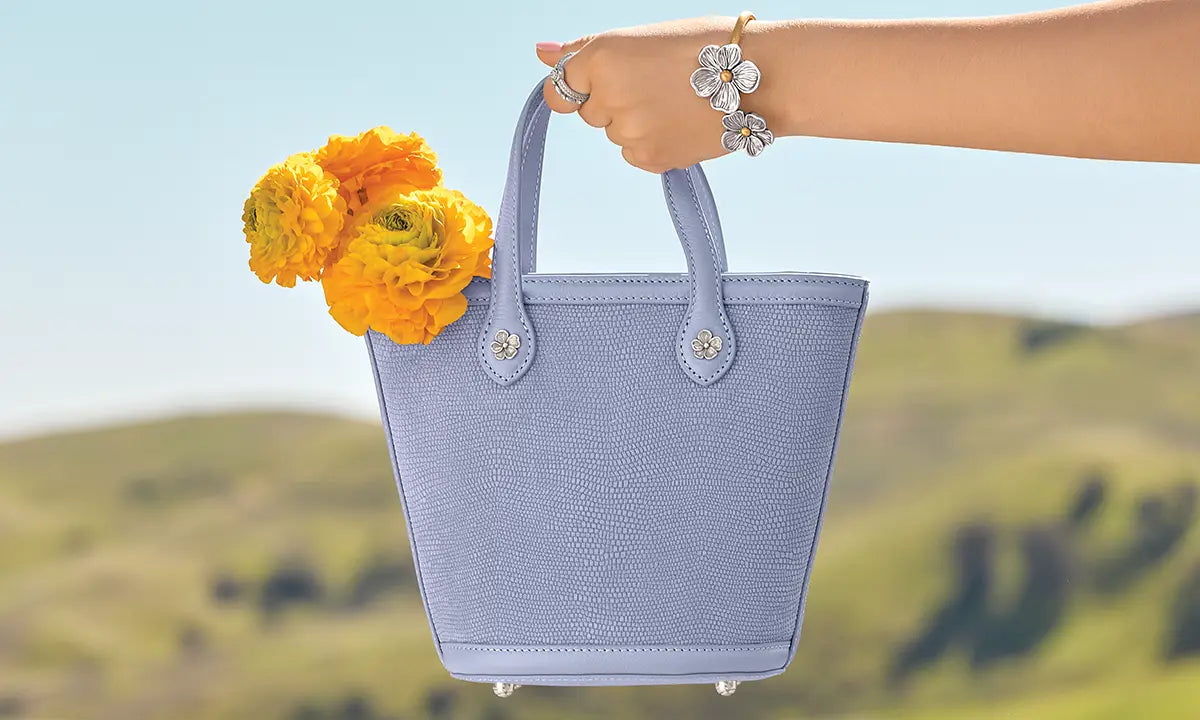 Light blue Brighton handbag with yellow flowers held by a hand against a blurred natural background