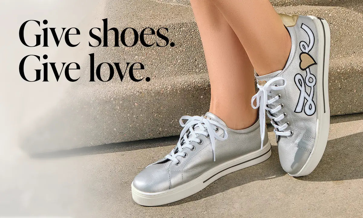 Silver sneakers with a heart and paw print design on a concrete surface, with text 'Give shoes. Give love.'