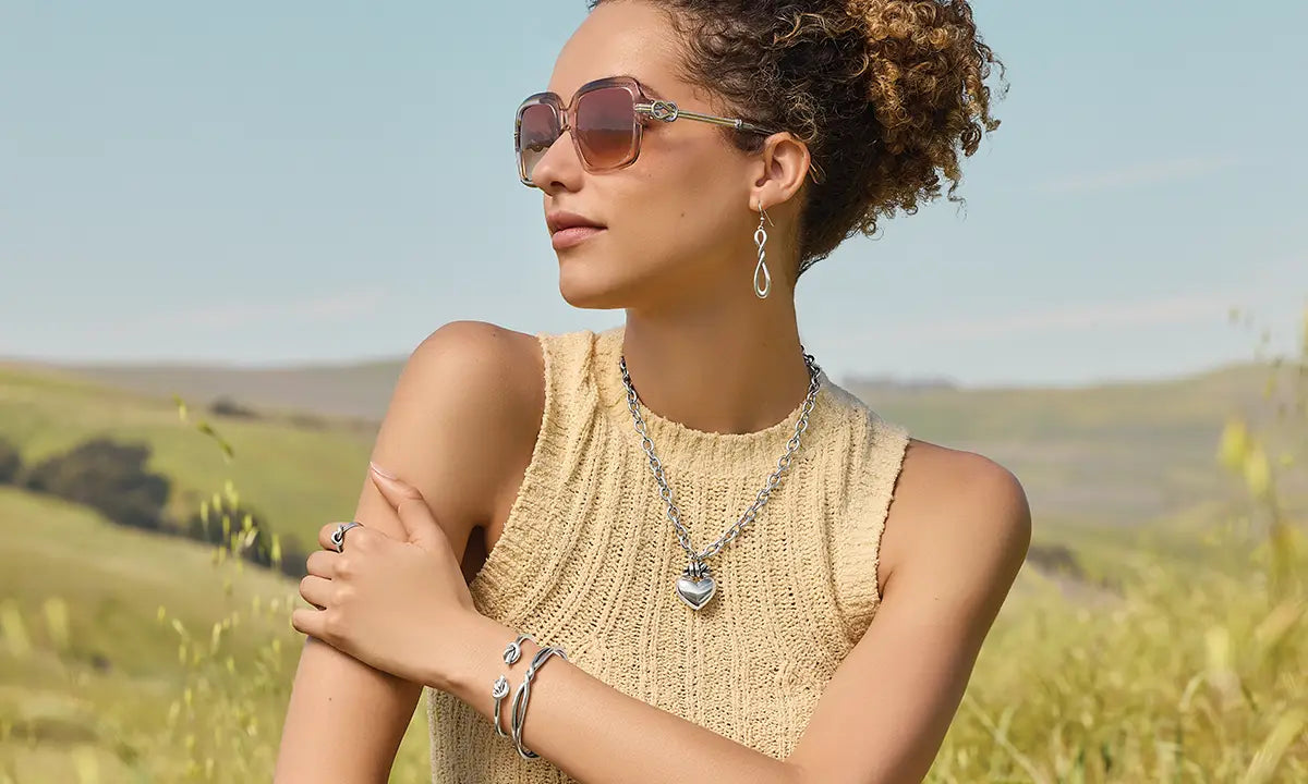 Woman wearing Brighton sunglasses and jewelry in a field with mountains in the background