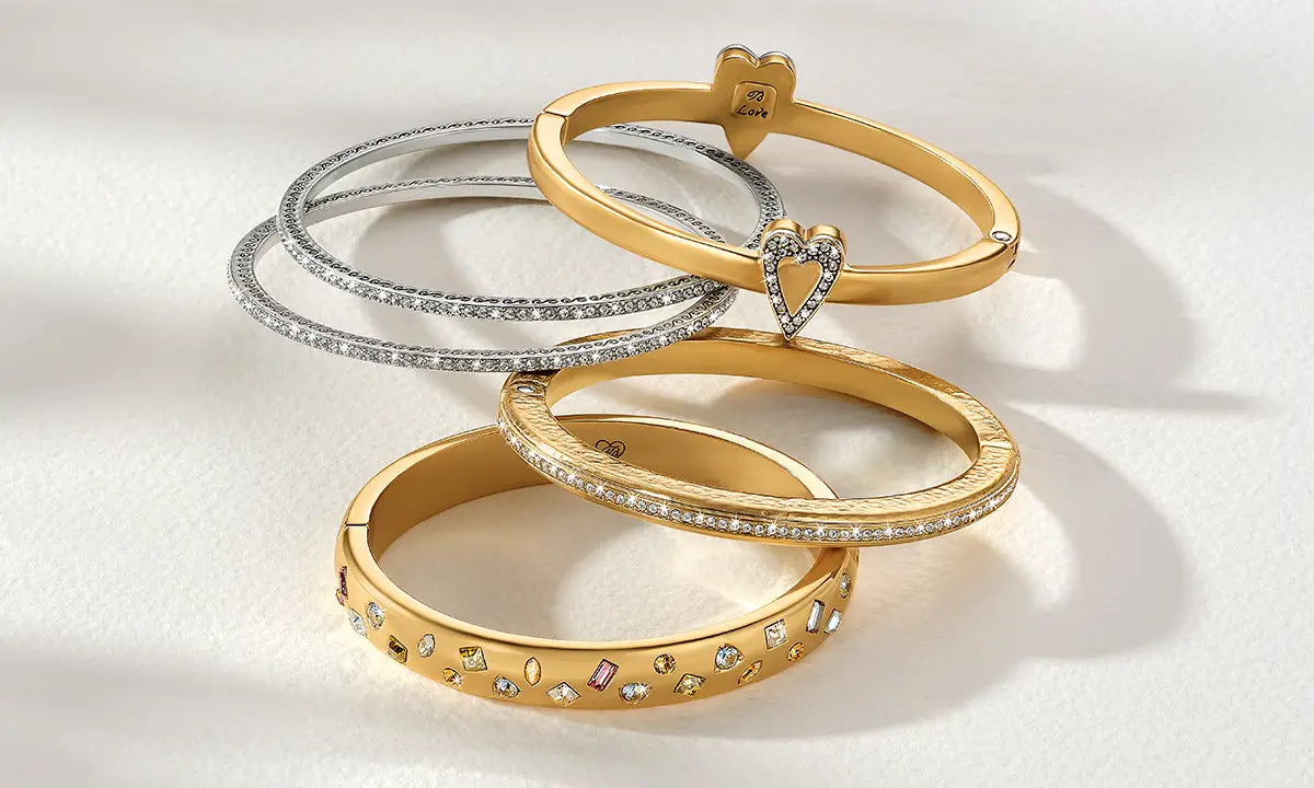 Four bangle bracelets arranged on a white surface, including two gold bangles, two silver bangles, and one gold bangle with small colored gemstones and a heart-shaped pavé detail.