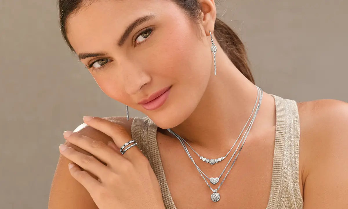 A close-up of a woman wearing layered silver necklaces with heart and oval pendants, matching drop earrings, and a silver ring, against a neutral beige background.