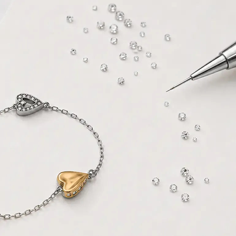 A silver chain bracelet with a gold heart charm and a small heart charm set with clear stones, displayed on a light background with scattered clear rhinestones and a metal pin.