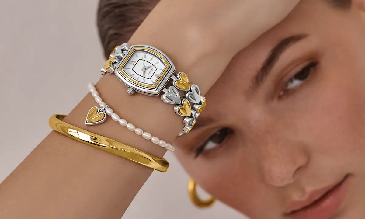 A close-up lifestyle shot of a woman's wrist adorned with a silver and gold watch, a gold bangle, a pearl bracelet with a heart charm, and a silver heart-link bracelet.