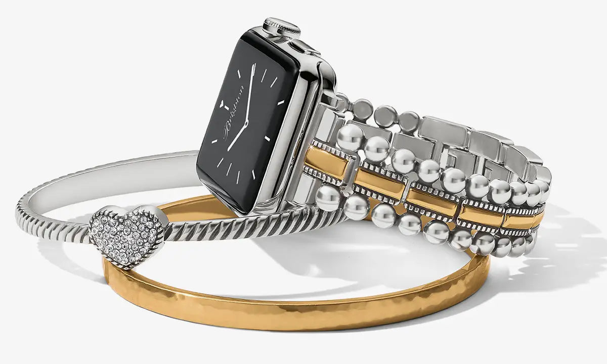 A silver-toned smartwatch with a rectangular black face and a multi-band bracelet featuring silver and gold elements, displayed on top of a gold bangle with a heart-shaped crystal charm against a light gray background.
