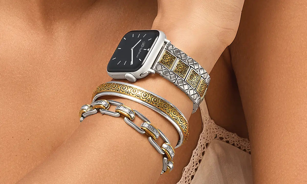 A close-up of a person's wrist wearing a silver smartwatch with a textured geometric link band, layered with two gold and silver bracelets.