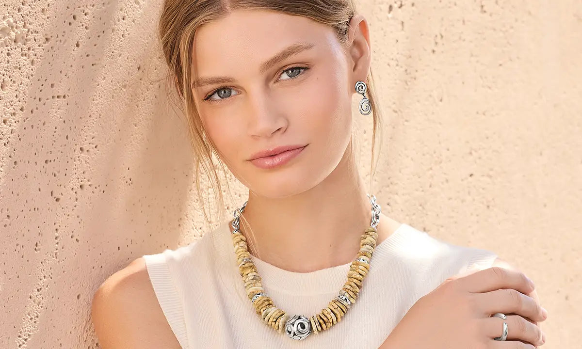 A close-up lifestyle shot of a model wearing a chunky gold and silver beaded necklace, matching drop earrings, and a ring, set against a textured beige stucco wall.