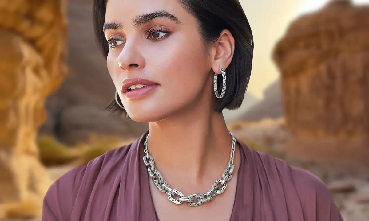 A woman wearing a chunky silver chain-link necklace and matching silver hoop earrings, photographed outdoors against a blurred desert rock background.