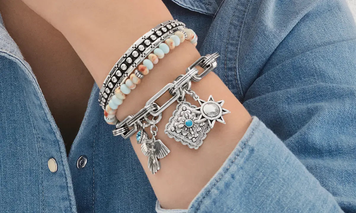 A close-up of a person's wrist wearing multiple bracelets, including silver charms, beaded bands, and a chain bracelet, paired with a blue denim shirt.