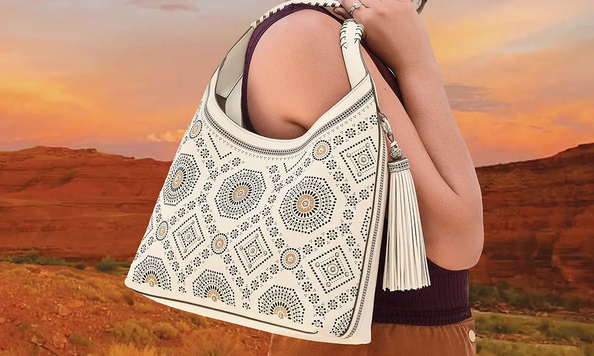 A person outdoors at sunset carries a large shoulder bag with a cream base and intricate geometric embroidery in black, blue, and tan. The bag features a tassel detail and is shown against a desert landscape.