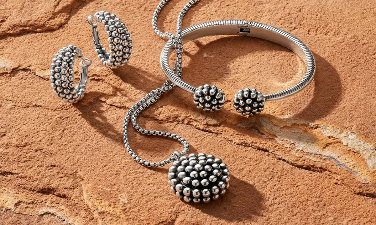 A silver jewelry set featuring a beaded ball pendant necklace, matching hoop earrings, and a beaded ball cuff bracelet, arranged on a textured orange-brown rock surface.