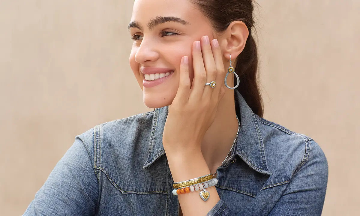 A woman wearing a silver hoop earring and a stack of beaded bracelets on her wrist, smiling and touching her face. She is wearing a denim shirt against a neutral beige background.
