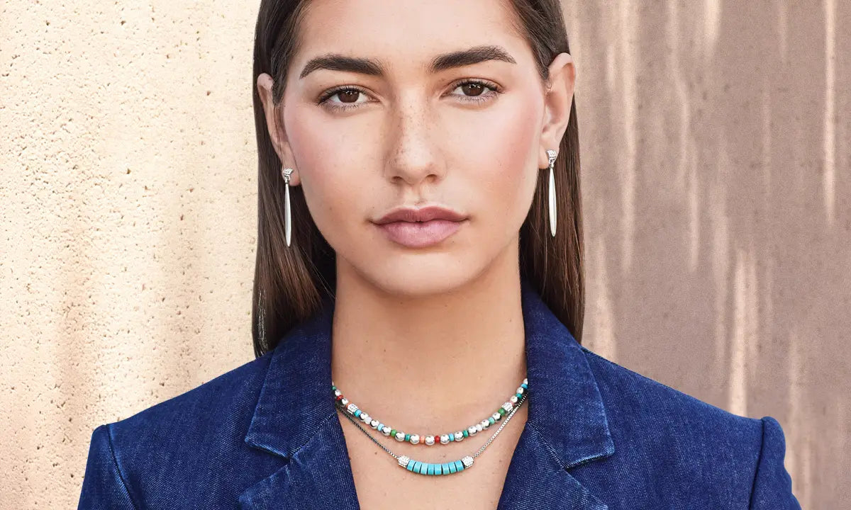 A woman wearing two necklaces, a turquoise beaded necklace and matching drop earrings, styled with a blue blazer against a textured beige wall.