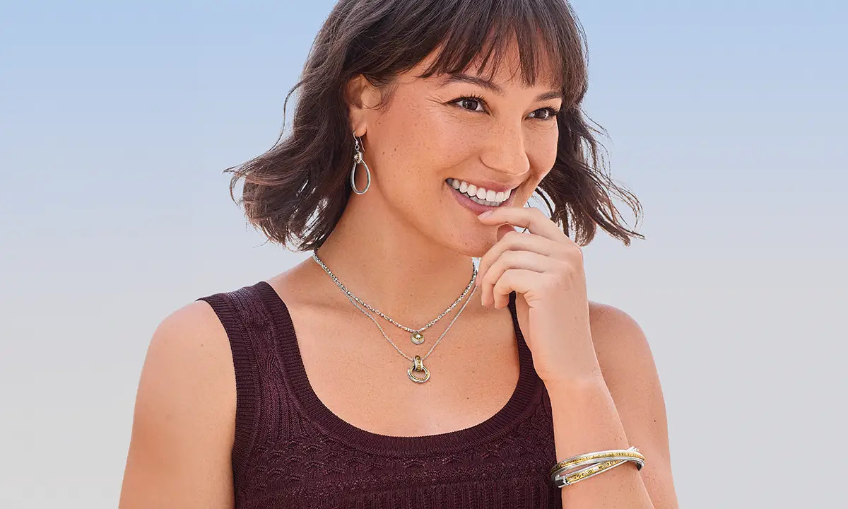 A smiling woman with short brown hair and bangs models layered silver necklaces, hoop earrings, and bracelets against a soft blue gradient background.