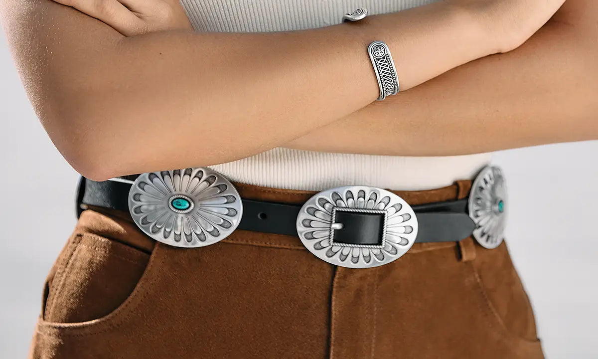 A close-up of a person wearing a black leather belt with three large silver concho-style buckles, one of which features a turquoise stone accent. The person is also wearing a silver woven cuff bracelet on their left wrist.