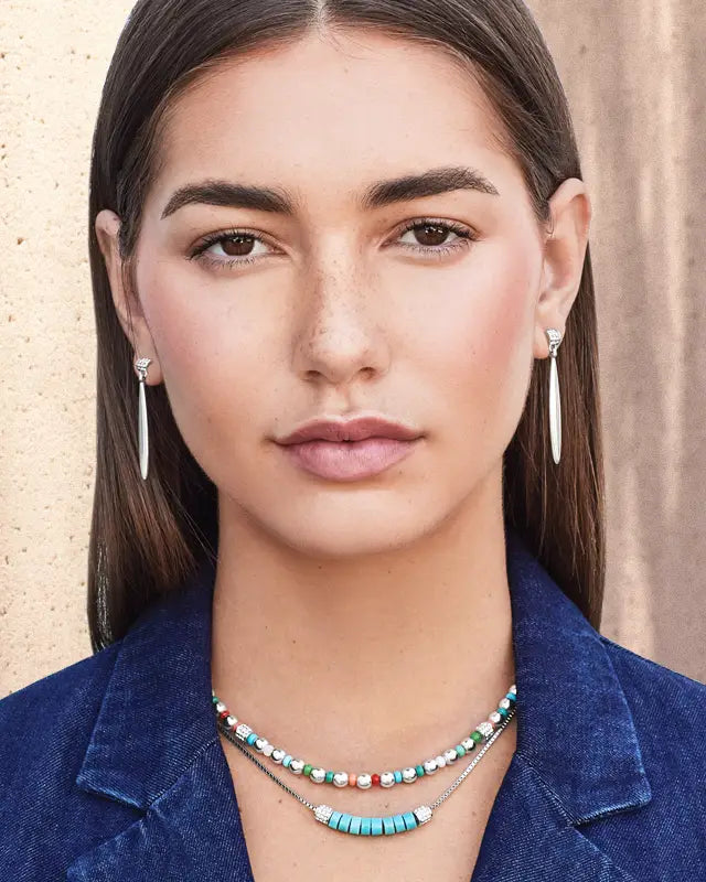A close-up portrait of a woman wearing a layered necklace and matching drop earrings. The necklace features turquoise, white, green, and red beads on silver-toned chains, paired with long silver drop earrings.