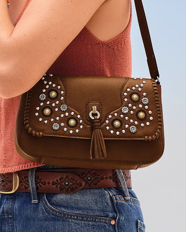 A brown leather crossbody bag with western-style silver and gold rhinestone embellishments, a tassel, and a braided strap, shown worn by a model in a rust-colored knit top and blue jeans.