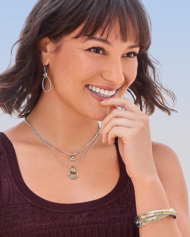 A smiling woman with short brown hair models layered silver and gold necklaces, hoop earrings, and bangles against a light blue sky background.