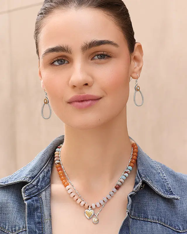 A close-up of a woman wearing a beaded necklace with a gold heart pendant and matching drop earrings, styled with a denim jacket against a neutral beige background.