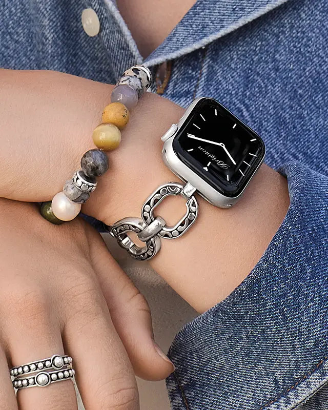 A close-up of a person's wrist wearing a silver chain-link watch strap attached to a smartwatch, alongside a beaded bracelet and rings, against a blue denim shirt background.