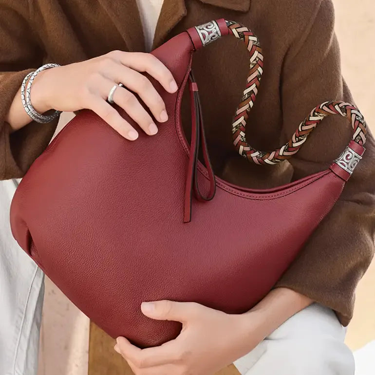 A person in a brown coat holds a large pebbled leather hobo bag in a deep burgundy red. The bag features a braided shoulder strap with red, beige, and white stripes and silver-toned metal hardware.