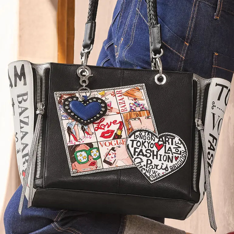 A black leather tote bag with silver zippers, braided shoulder straps, and decorative patches, shown being worn by a person in blue jeans. The bag features a central patch collage with a blue heart charm and a black-and-white heart patch.