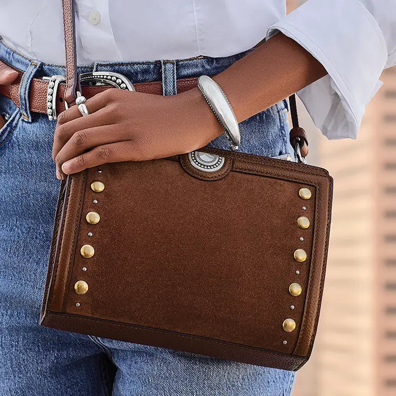 A brown suede crossbody handbag with gold-tone studs and silver-toned western-style hardware, worn across a person's torso over blue jeans and a white shirt.