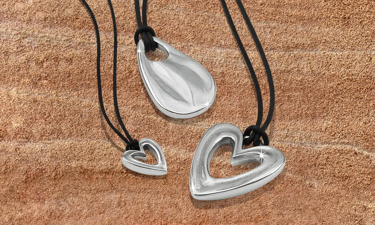 Three silver heart-shaped pendant necklaces with black cords, displayed on a textured brown surface.
