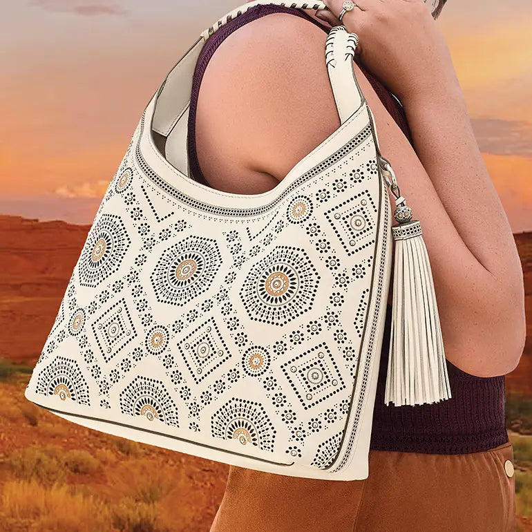 A woman carries a large white shoulder bag with bohemian-style geometric embroidery in black, gold, and brown. The bag features a tassel detail and is shown against a warm desert sunset background.