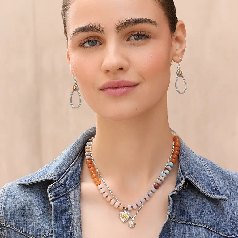 A close-up of a woman wearing a beaded necklace with a gold heart pendant and matching drop earrings, paired with a denim jacket against a soft beige background.