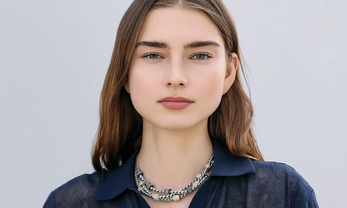 A close-up portrait of a woman with brown hair wearing a dark navy blue shirt and a chunky silver chain necklace with pave accents, set against a solid light gray background.