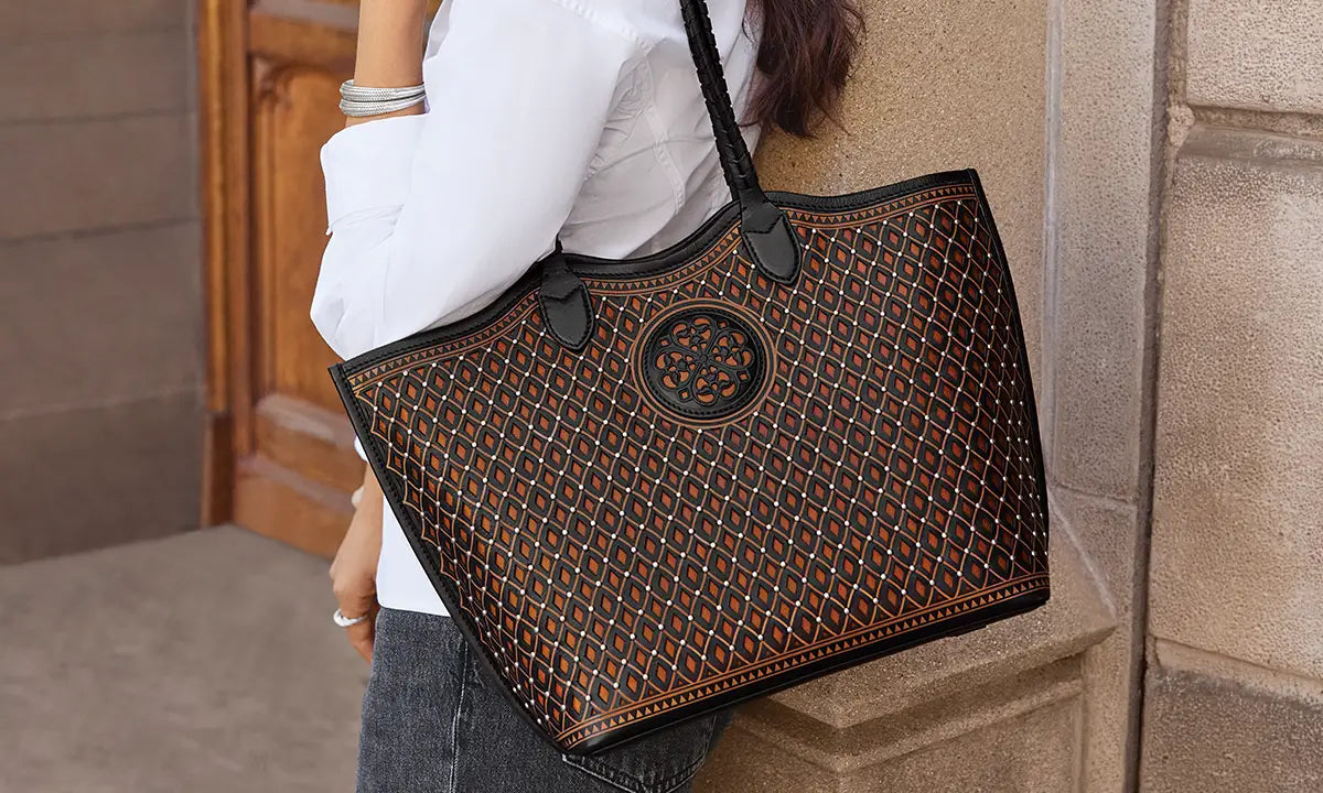 A person wearing a white shirt and dark jeans carries a large black tote bag with an intricate brown and orange geometric pattern and a circular medallion logo on the front, set against a stone wall background.