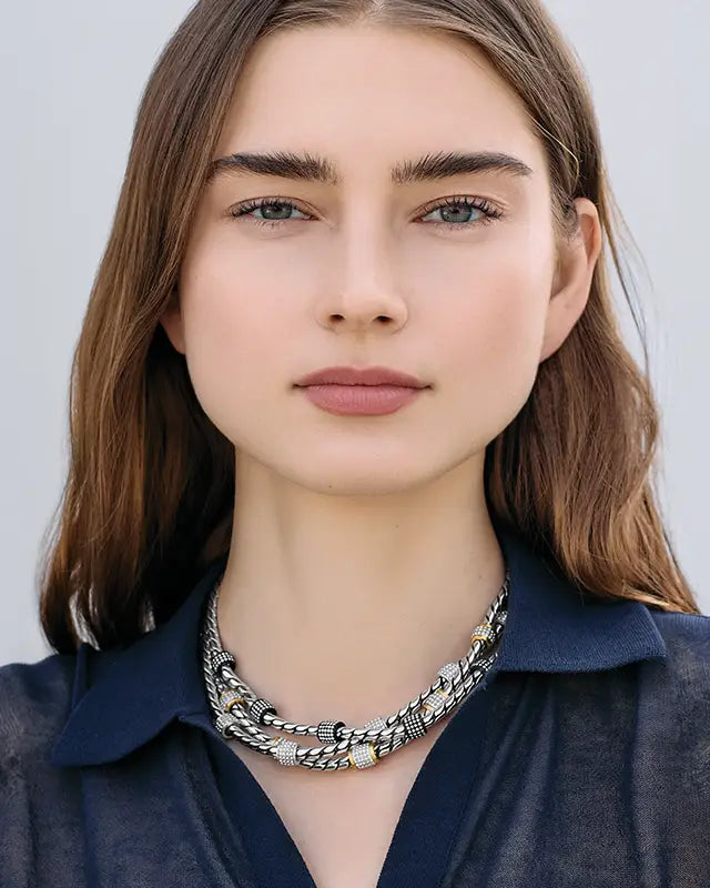 A close-up portrait of a woman with brown hair wearing silver collar necklaces and a dark navy collared shirt against a light gray background.