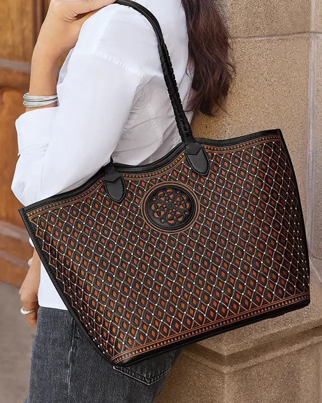 A woman wearing a white blouse and gray jeans carries a large black woven tote bag with a geometric diamond pattern and a circular medallion detail on the front, leaning against a stone wall.