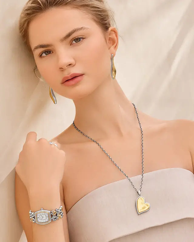 A woman models a silver chain necklace with a gold heart pendant, matching gold and silver earrings, and a silver watch with a crystal-encrusted face, set against a draped beige background.