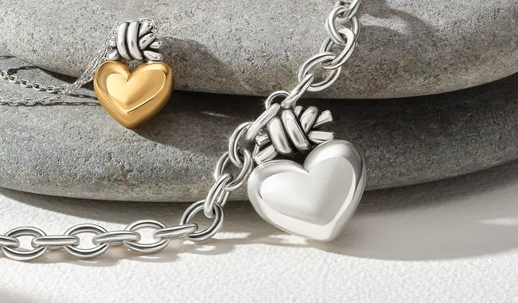 A close-up of a silver chain necklace with a heart-shaped pendant, resting on stacked gray stones. A second matching necklace with a gold heart pendant is partially visible in the background.