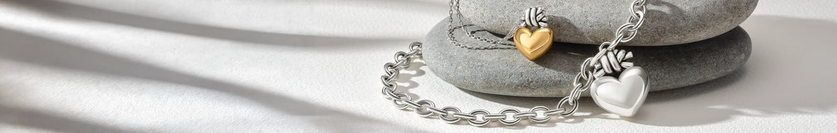 Silver chain necklaces with heart charms, including a gold heart and a silver heart, displayed on stacked gray stones with soft shadows.