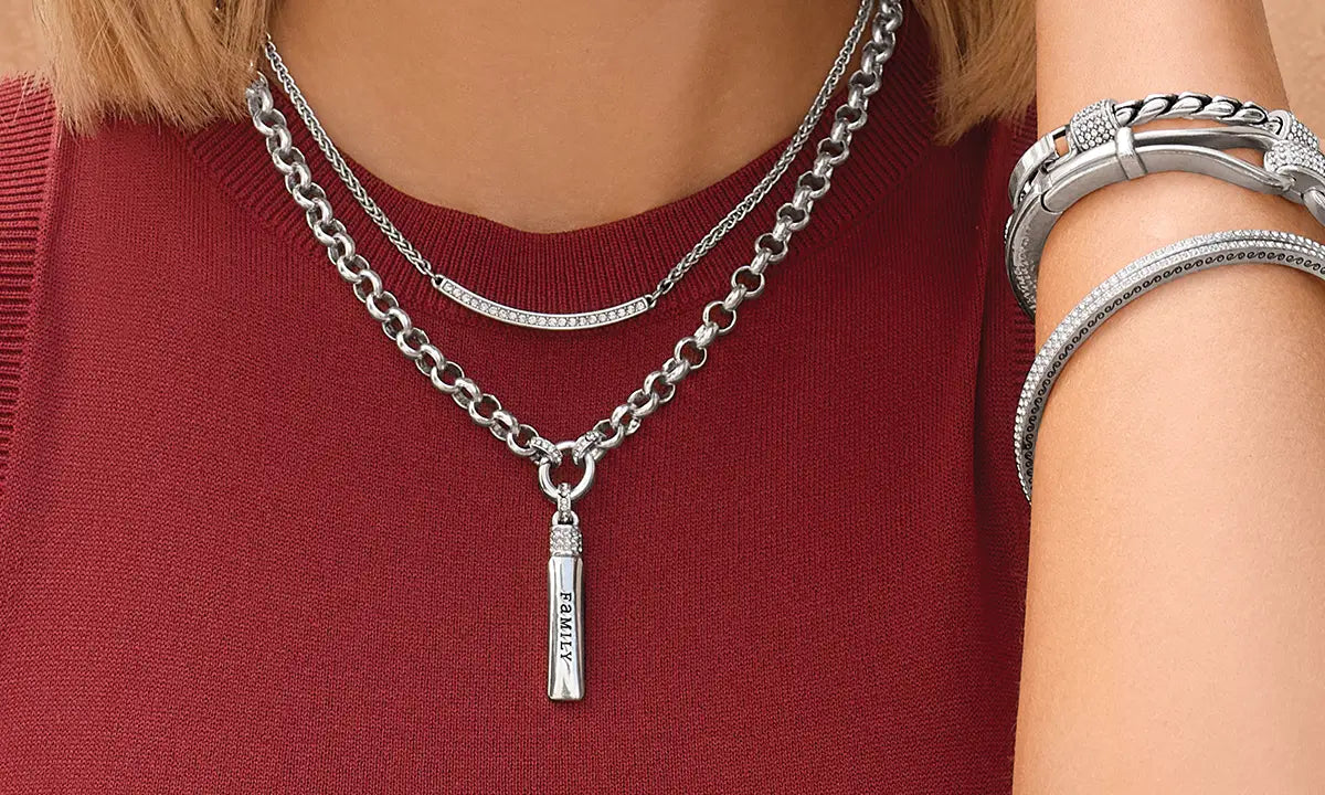 A close-up of a person wearing layered silver necklaces, including a vertical pencil-shaped pendant engraved with 'FAMILY', along with silver bracelets on the wrist, set against a dark red knit top.