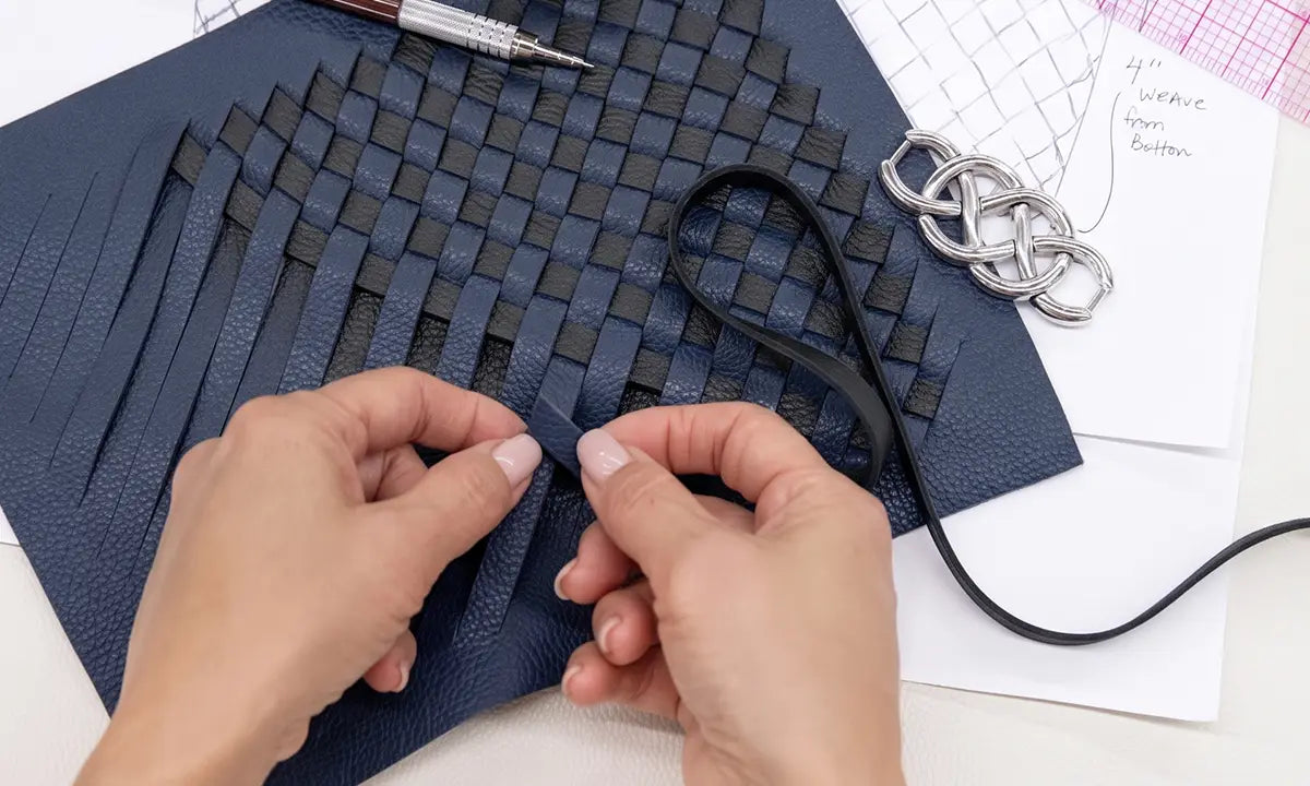 Hands weaving a dark navy blue leather strap over a matching textured leather sheet, with a pen, metal ring, and sketch notes visible on a white surface.