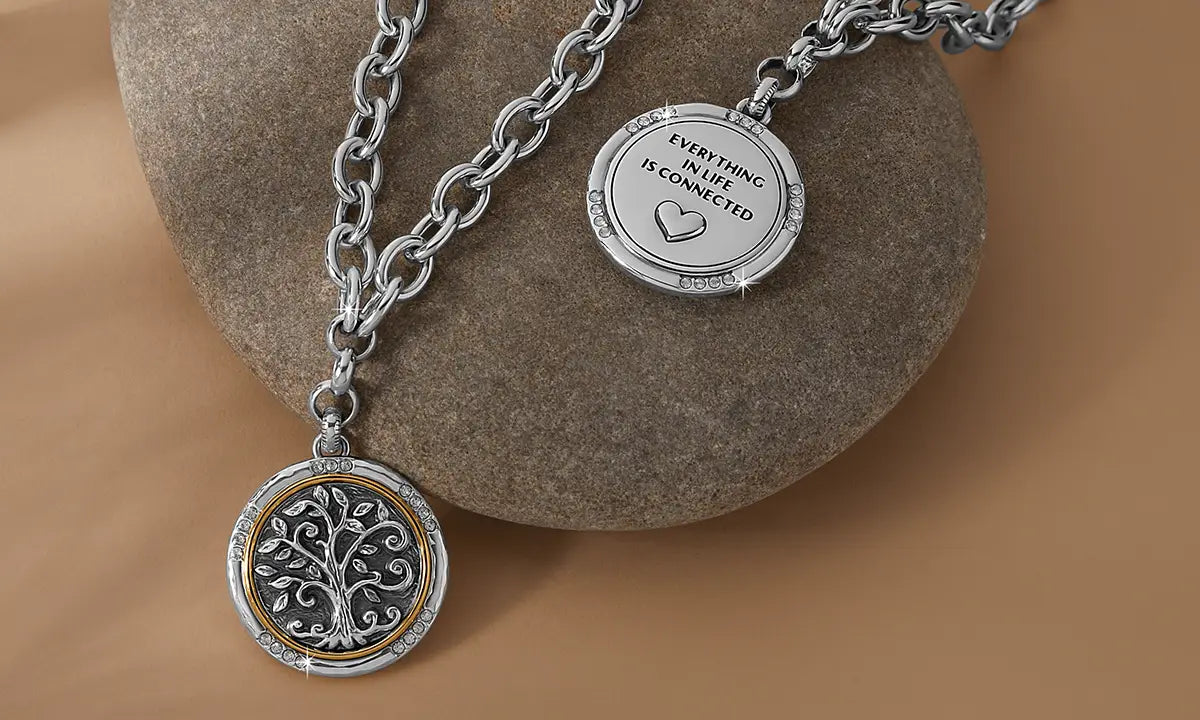 A silver-toned chain necklace displayed on a smooth stone, showing two of the same round medallion pendants. One pendant shows the back which is engraved with 'EVERYTHING ISN LIFE IS CONNECTED' and a heart, while the other shows the back, hanging below with a tree design in a silver and gold-tone center.