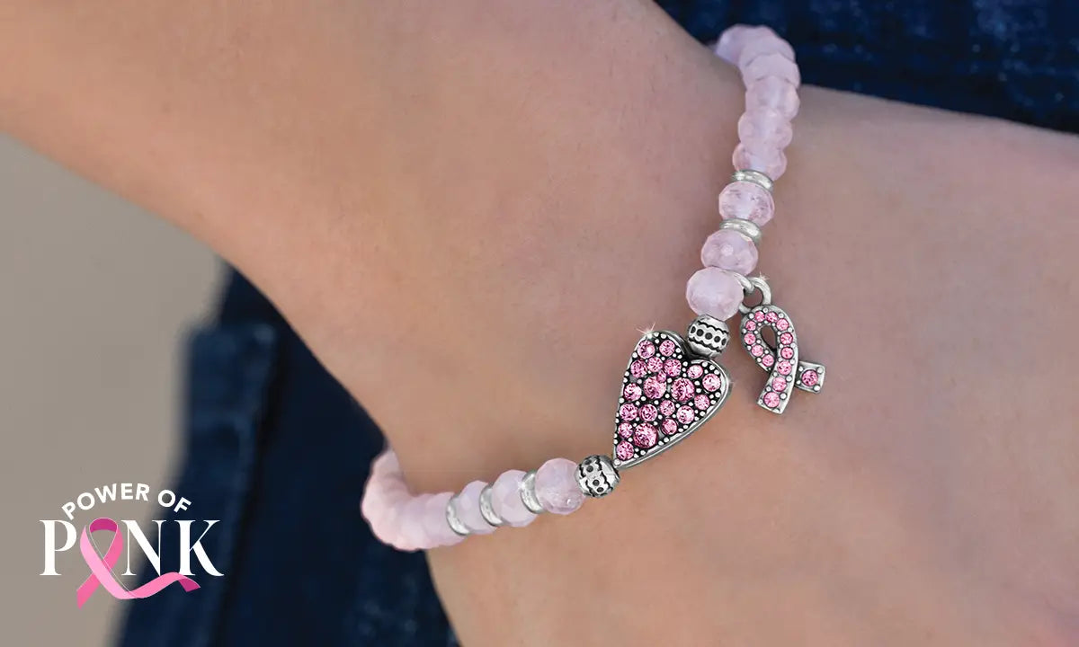 A close-up of a pink beaded bracelet worn on a wrist, featuring a heart-shaped charm paved with pink stones and a pink ribbon charm. The brand logo 'POWER OF PINK' is visible in the bottom-left corner.