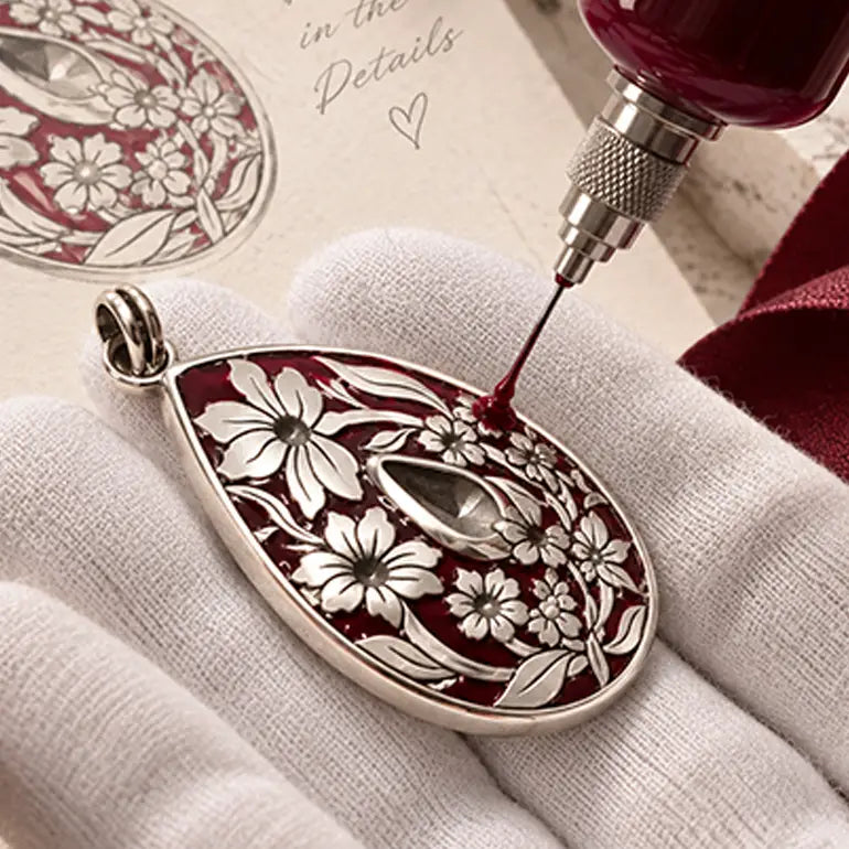 A close-up of a teardrop-shaped pendant with a silver-tone floral design over a deep red enamel background, being filled with dark red enamel by a jeweler's dropper. The pendant is held on a white gloved hand, with a matching inset detail view in the top-left corner.