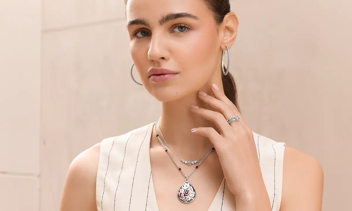 A woman wearing a silver-toned necklace with a teardrop-shaped pendant featuring red and white gemstone detailing, along with matching hoop earrings and a ring, posing against a neutral beige background.