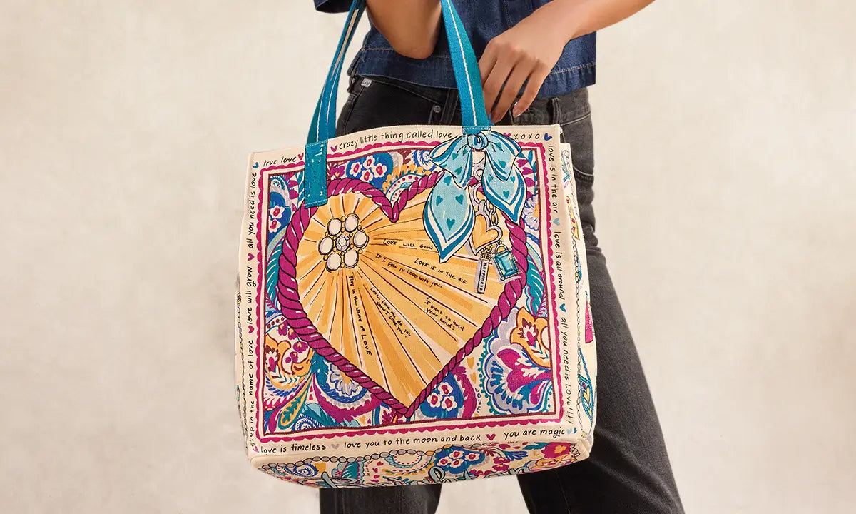 A person wearing dark jeans and a blue denim top holds up a large tote bag decorated with a colorful hand-painted style heart design, floral motifs, and script text, against a plain beige wall.