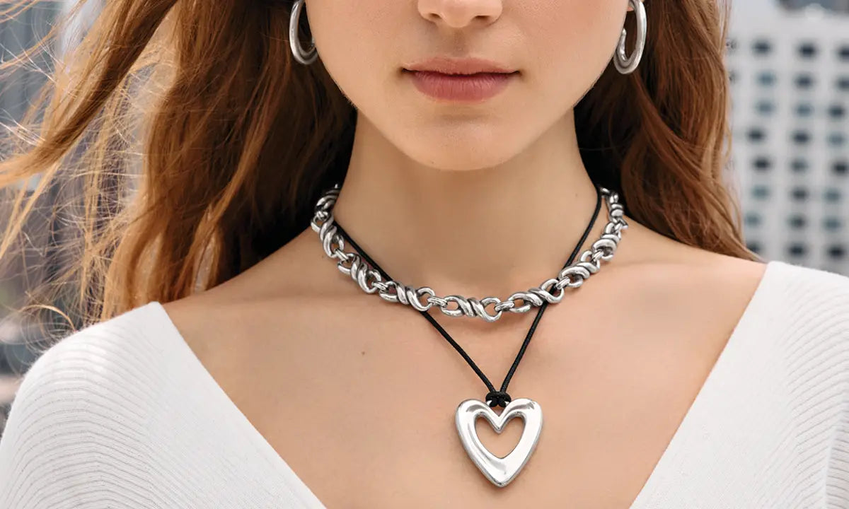 A close-up of a woman wearing a layered necklace set featuring a chunky silver chain choker and a black cord necklace with a polished silver open heart pendant.