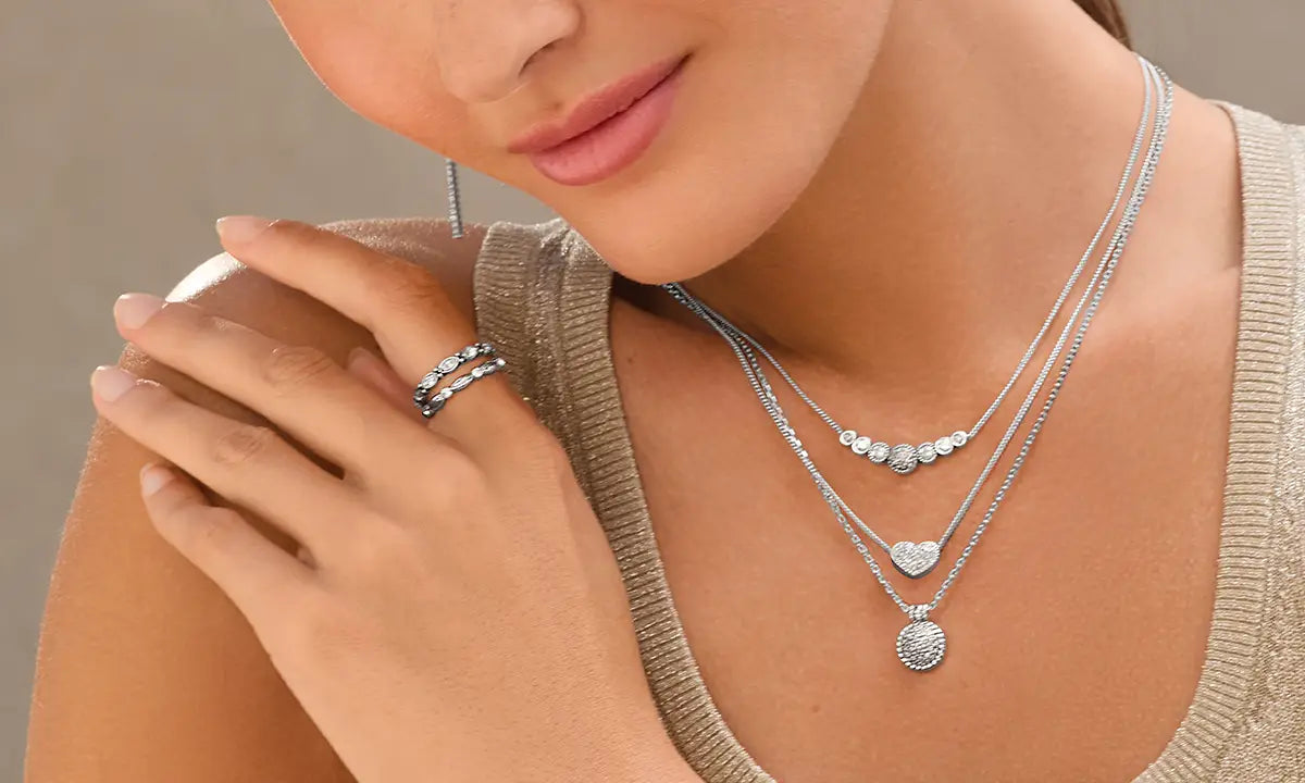 A close-up lifestyle shot of a model wearing a layered silver necklace set with a heart and oval pendant, along with a matching silver ring.