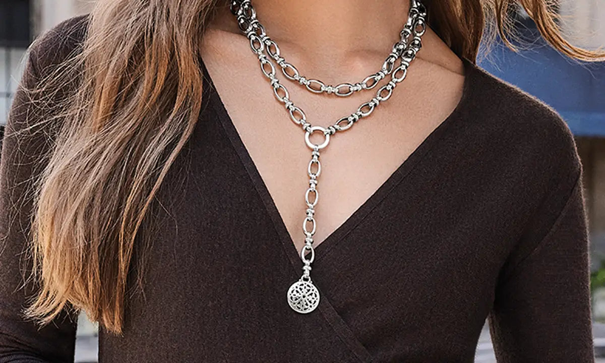A close-up of a woman wearing a layered silver chain necklace with a long Y-shaped drop and a circular pendant, paired with a dark brown V-neck top.
