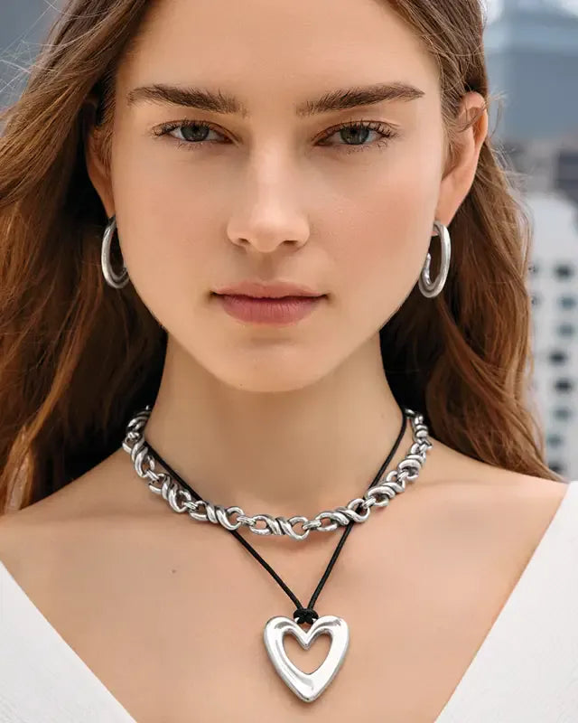 A close-up of a woman wearing a layered silver necklace with a heart-shaped pendant, a chunky silver chain choker, and silver hoop earrings.